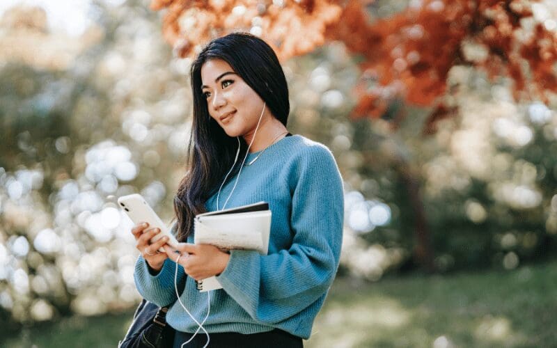 A woman stood under a tree listening to music on her mobile phone through headphones.